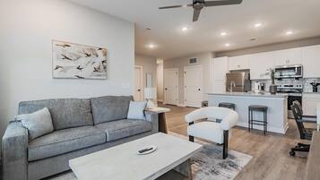 A modern living room with a grey sofa, a white armchair, and a coffee table. A ceiling fan is visible, and the room has a neutral color palette.