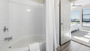 Un bagno moderno con pareti piastrellate di bianco, una vasca freestanding e un'apertura che conduce a una camera da letto con una grande finestra.