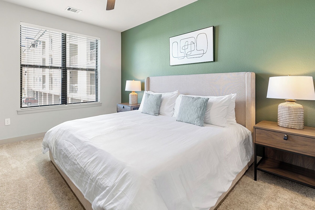 A bedroom with a large bed, two bedside tables with lamps, a window with blinds, and a green accent wall.