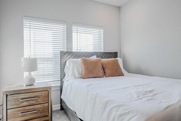 Une chambre à coucher comprenant un lit, deux fenêtres équipées de stores, une table de chevet avec une lampe et une commode.
