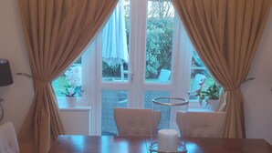 Un comedor con candelabro, cortinas color crema, una mesa con sillas y vistas a zonas verdes al exterior.