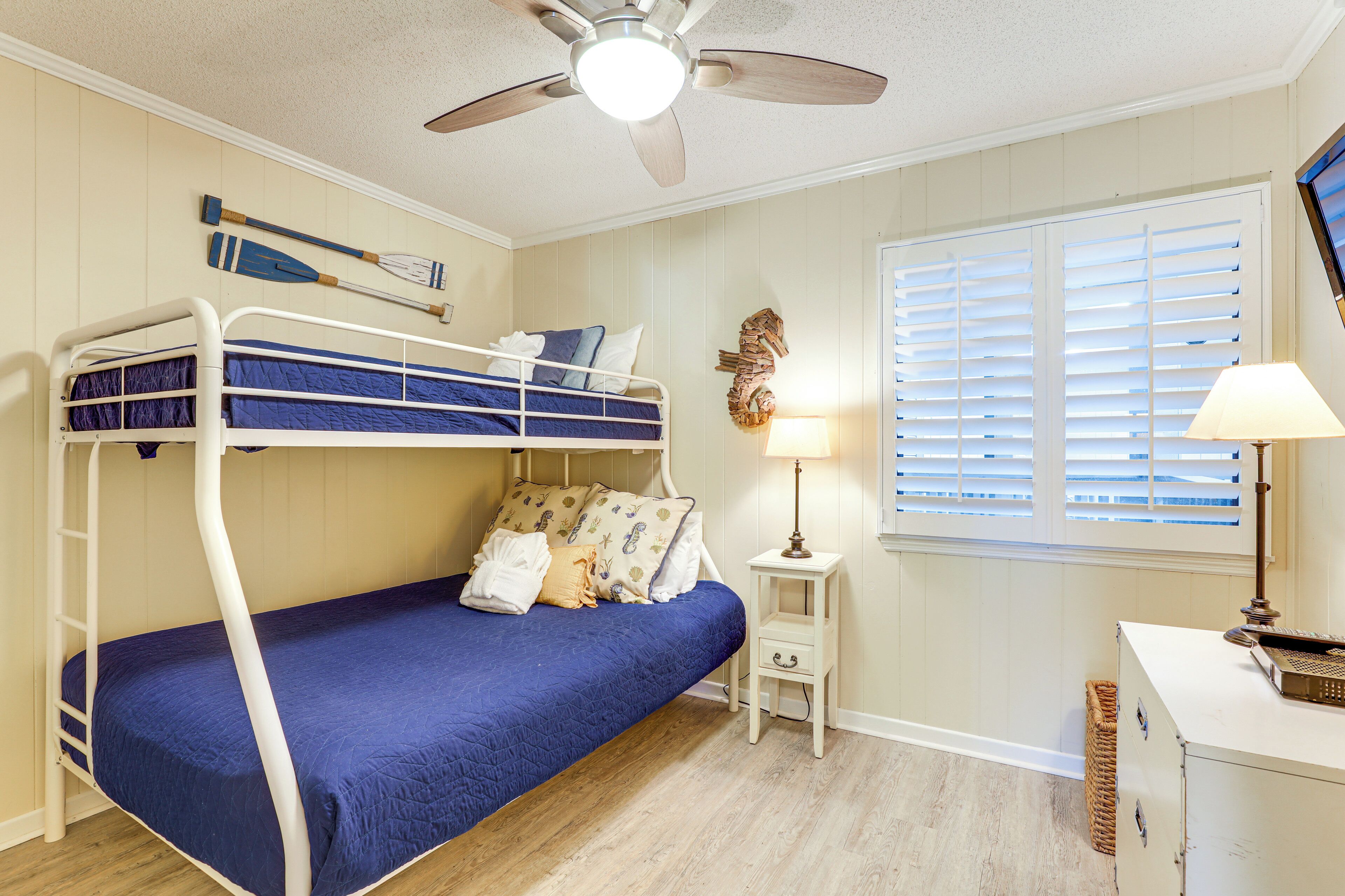 A bunk bed room with a ceiling fan, a small bedside table with a lamp, and a window with blinds.