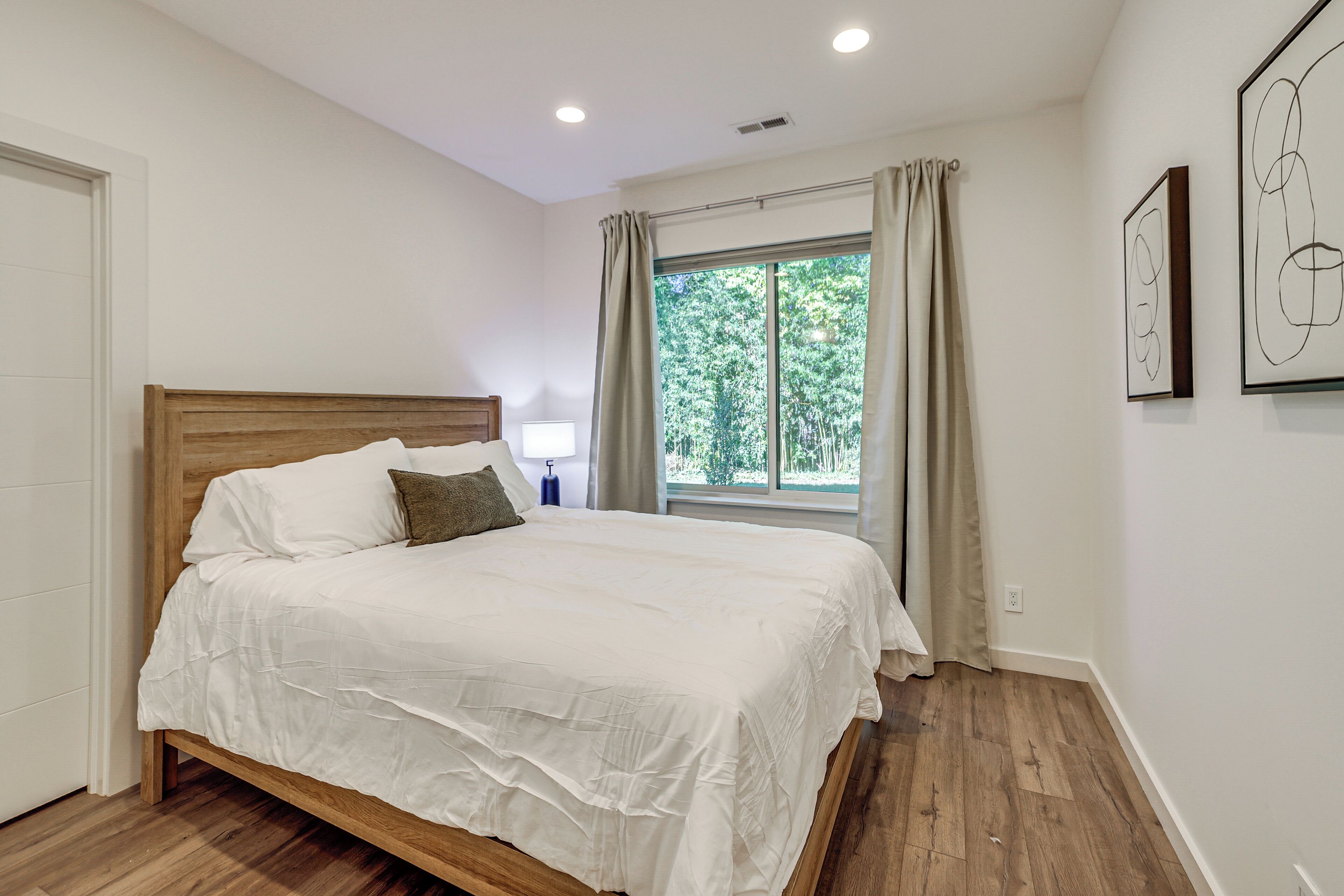 Ein Schlafzimmer mit einem hölzernen Bett, einem Fenster mit Vorhängen und zwei gerahmten abstrakten Kunstwerken an der Wand.