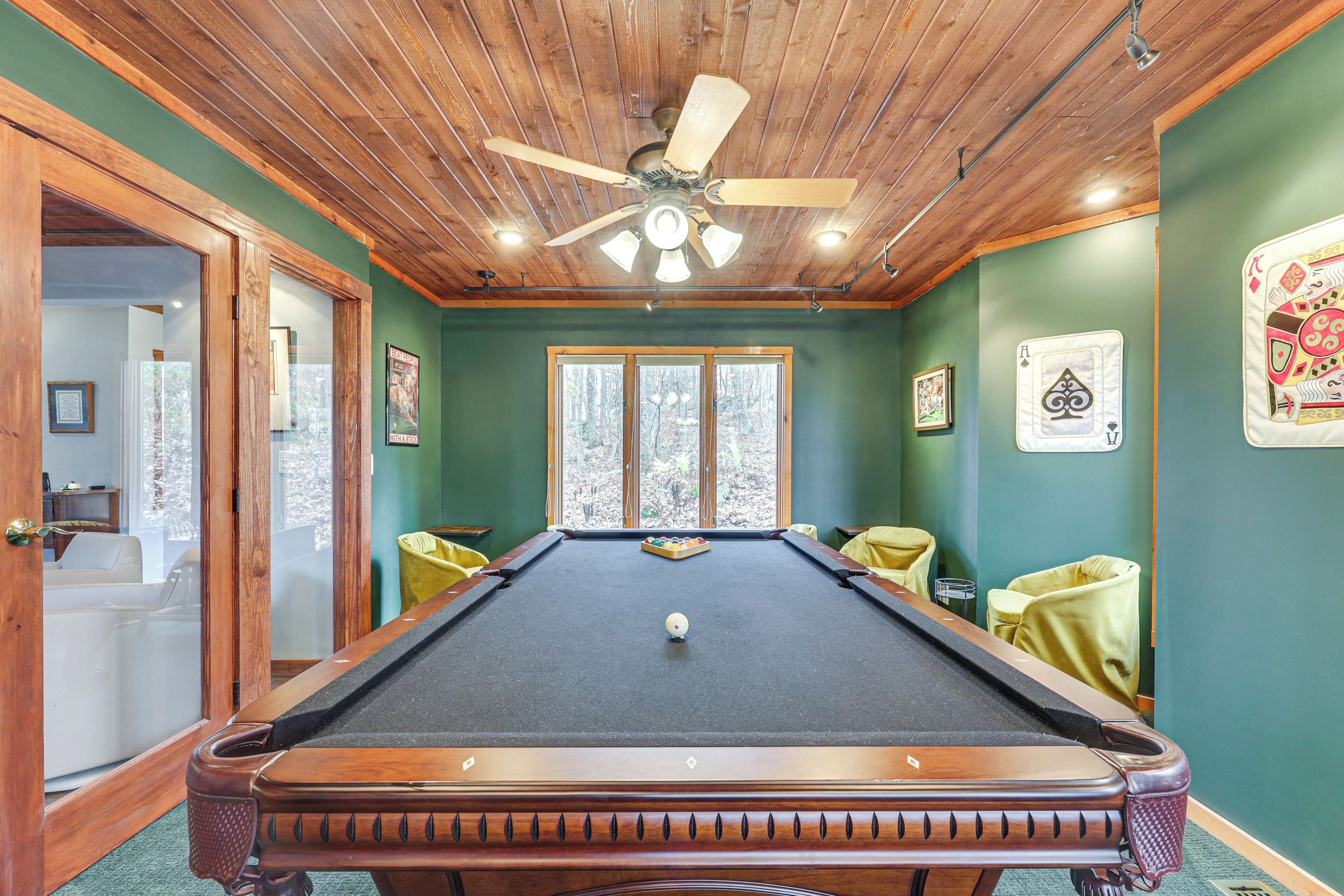 A pool table with a ceiling fan, green walls, and a window with a view of trees.