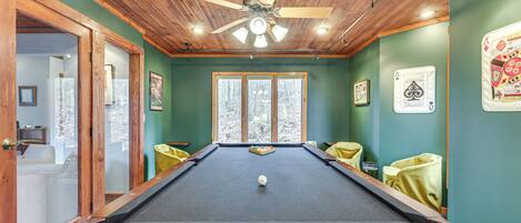 A pool table with a ceiling fan, green walls, and a window with a view of trees.