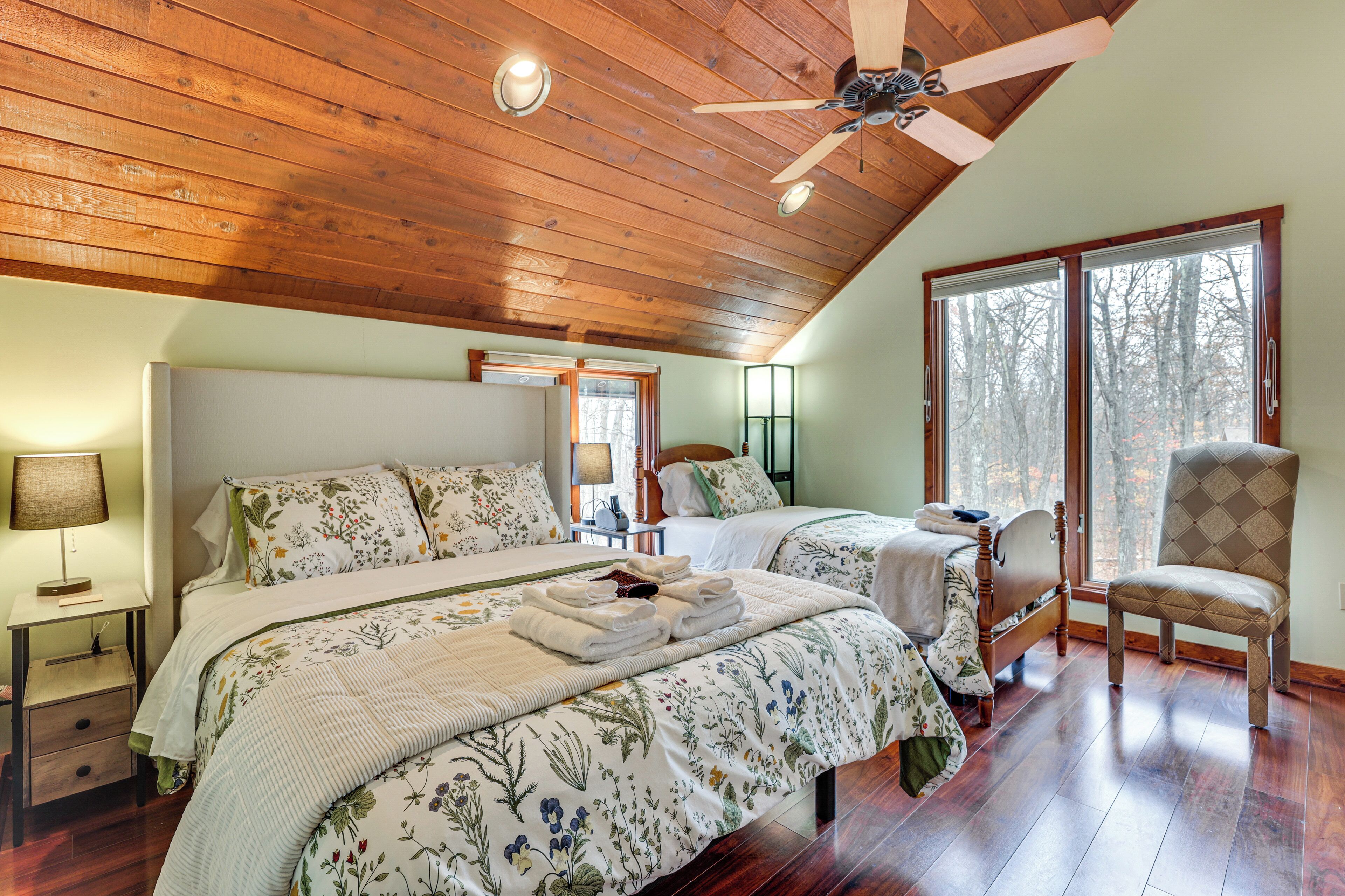 A bedroom with two beds, wooden ceiling, large windows, and a chair.
