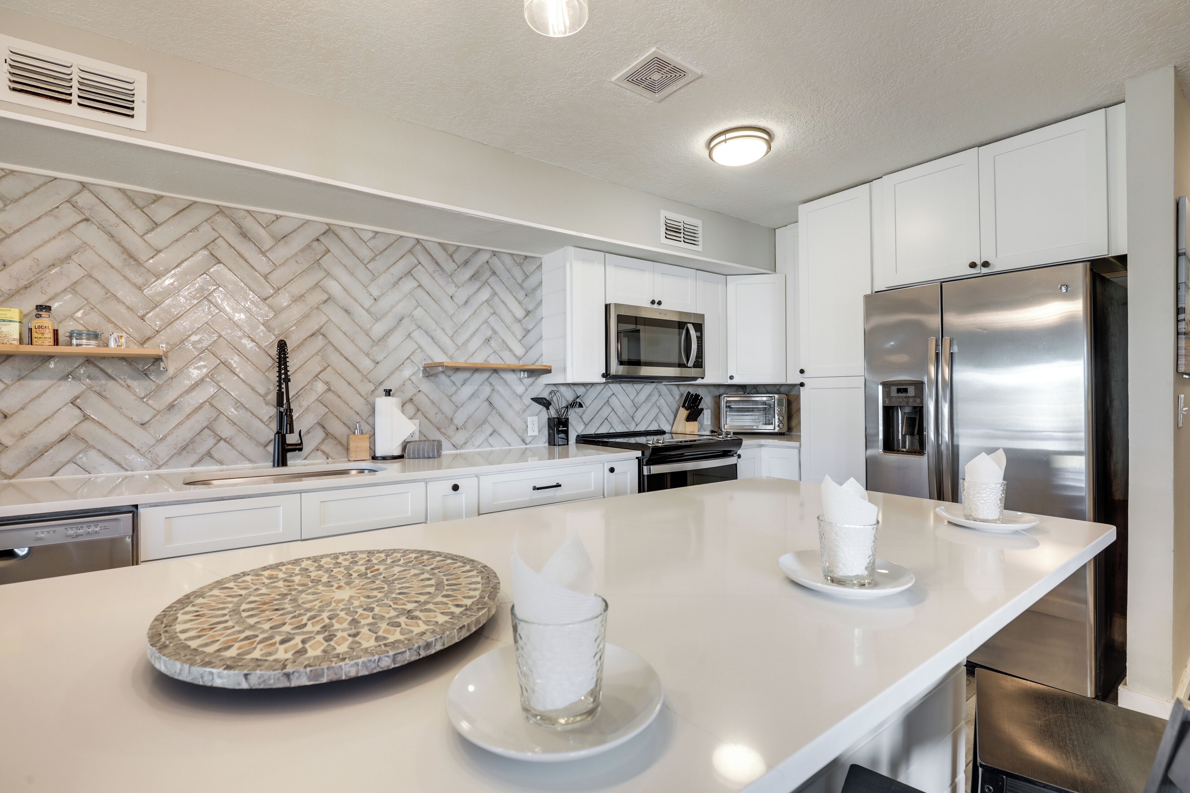 Une cuisine moderne avec des meubles blancs, un dosseret à motifs en chevrons, des appareils en acier inoxydable et un îlot central agrémenté d’une assiette décorative et de porte-serviettes.