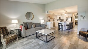 Un salon moderne comprenant un canapé, une table basse et des fauteuils, un coin cuisine avec un ensemble de salle à manger, et un couloir menant à une chambre.