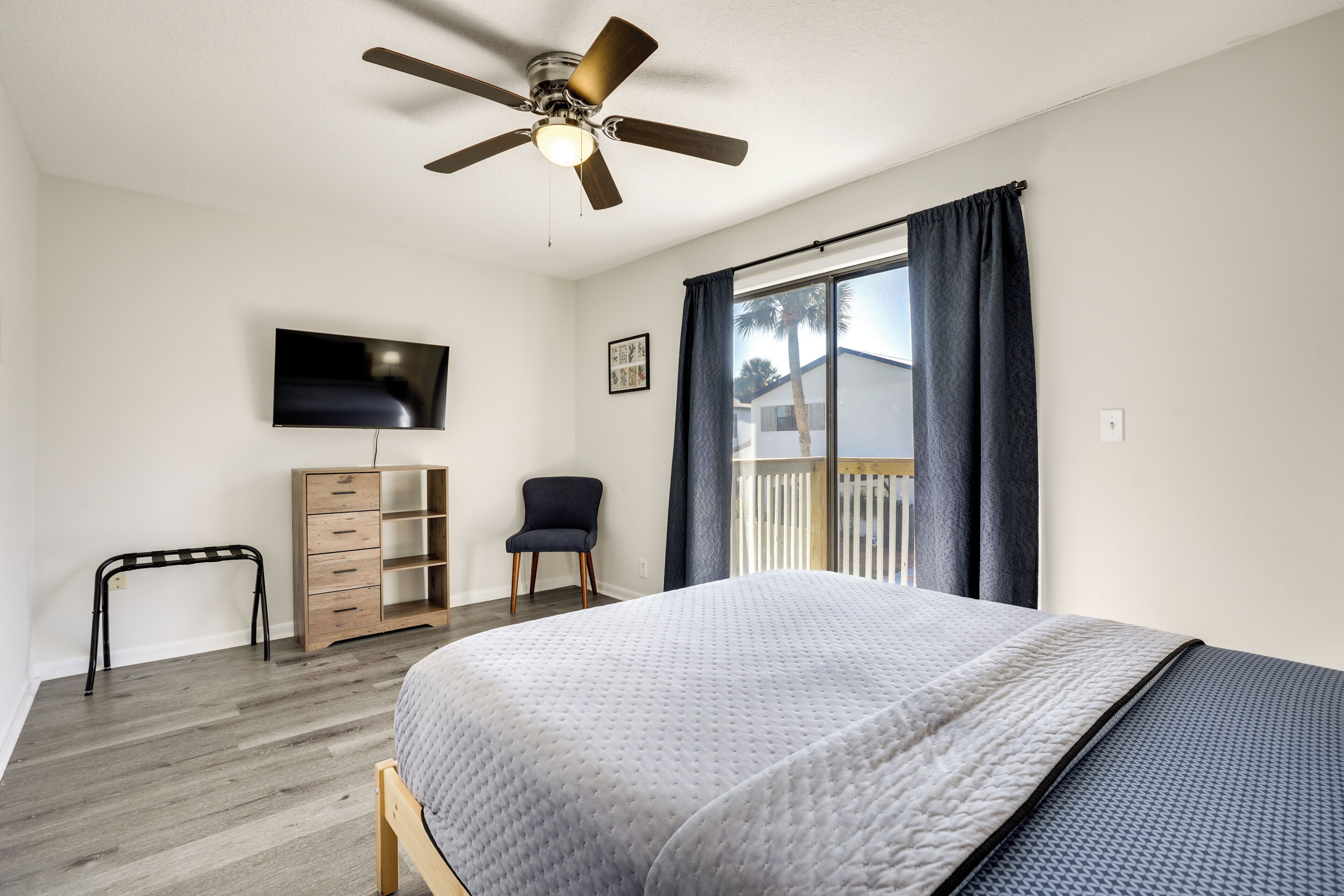 Quarto com cama, ventoinha no teto, televisão, cadeira, escrivaninha e janela com cortinas.