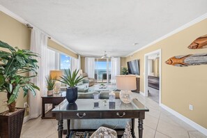A spacious living area with a glass coffee table, a sofa, and a TV. There are plants and a lamp on a side table. - 701 Twin Towers - Daytona Beach Oceanfront Vacations, Inc (Daytona Beach Shores)