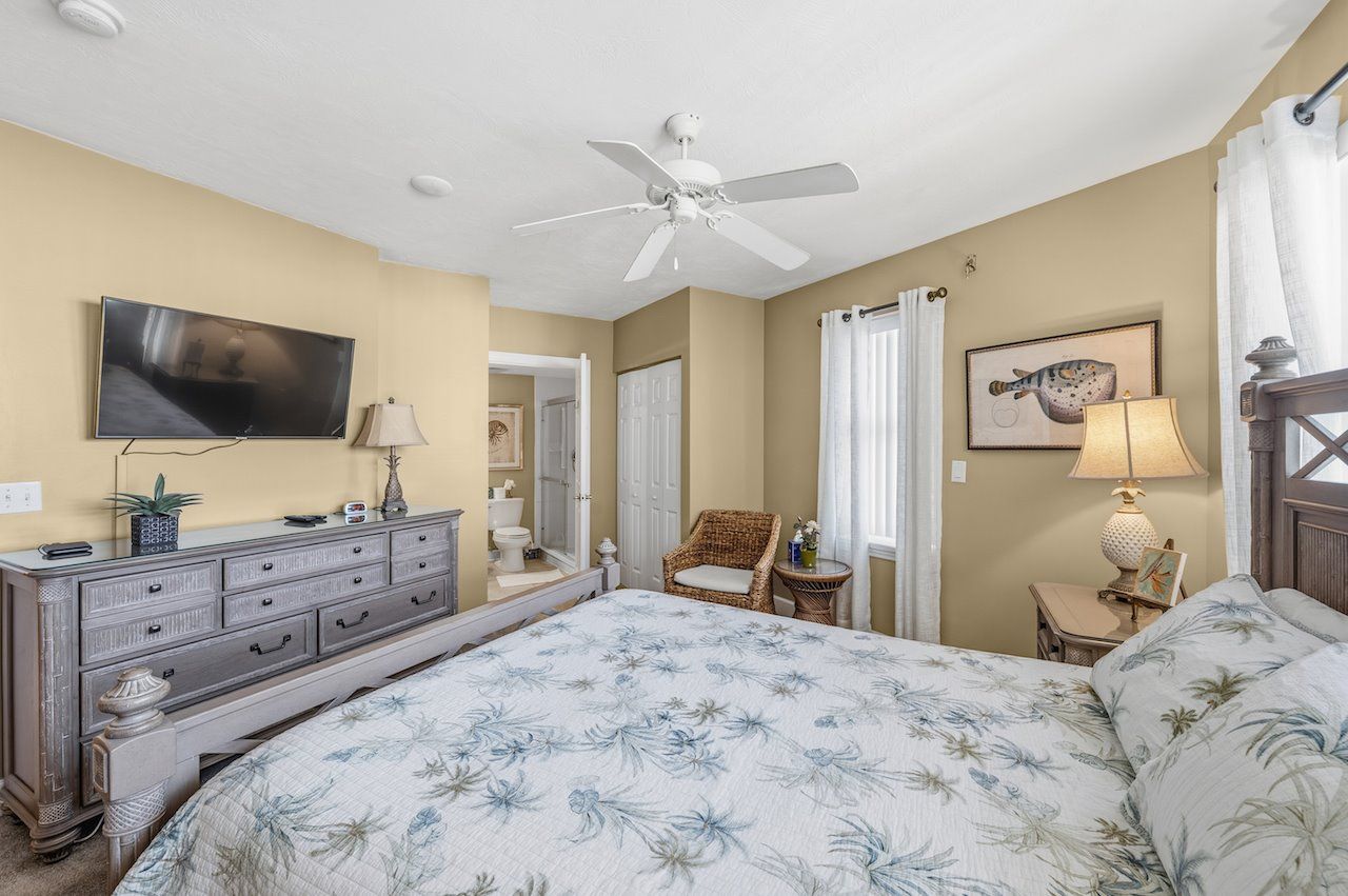 A bedroom with a bed, dresser, TV, ceiling fan, and a door leading to another room.
