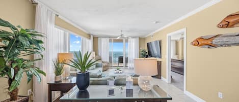 A spacious living area with a glass coffee table, a sofa, and a TV. There are plants and a lamp on a side table.