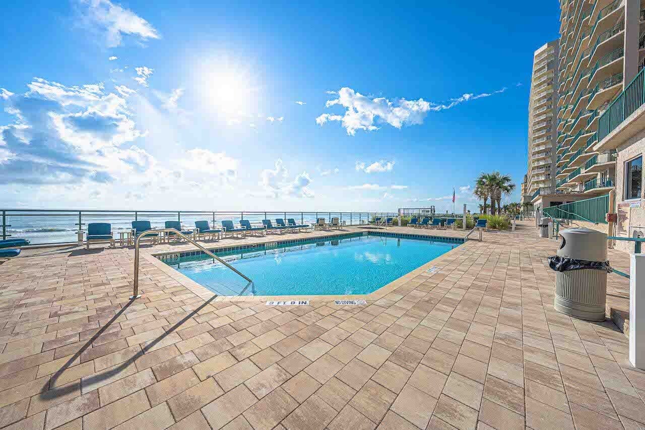 A pool area with a view of the ocean, lounge chairs, and a high-rise building in the background.