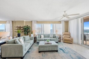 Un salon spacieux, doté d’un canapé de couleur claire, d’un pouf assorti et d’un ventilateur de plafond. On y trouve deux petites tables d’appoint avec des lampes et des objets décoratifs. Une grande fenêtre offre une vue sur le panorama urbain.