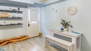 Ein Zimmer mit Holzboden, weißen Wänden, einer weißen Tür, Regalen mit Glaswaren, einer Uhr und einem kleinen Tisch mit einer Pflanze.