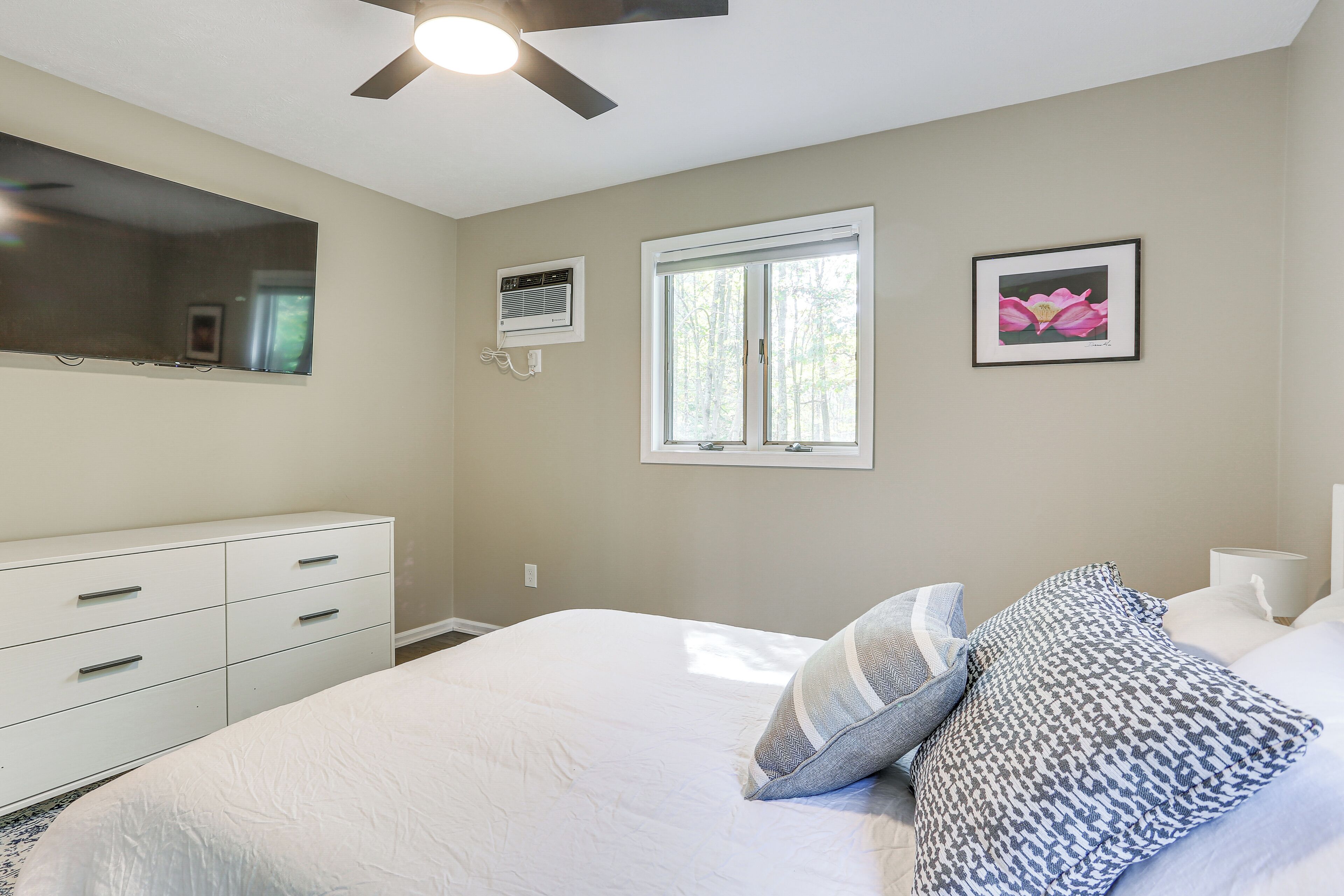 Un dormitorio con cama, cómoda, ventilador de techo, ventana y un cuadro en la pared.