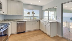 Een moderne keuken met witte kasten, een RVS vaatwasser en een groot raam met uitzicht op een waterpartij met palmbomen.