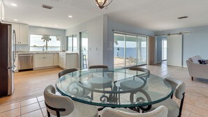 Een ruime woonkamer met een glazen eettafel en stoelen, een keuken met witte kasten, en uitzicht op het strand.