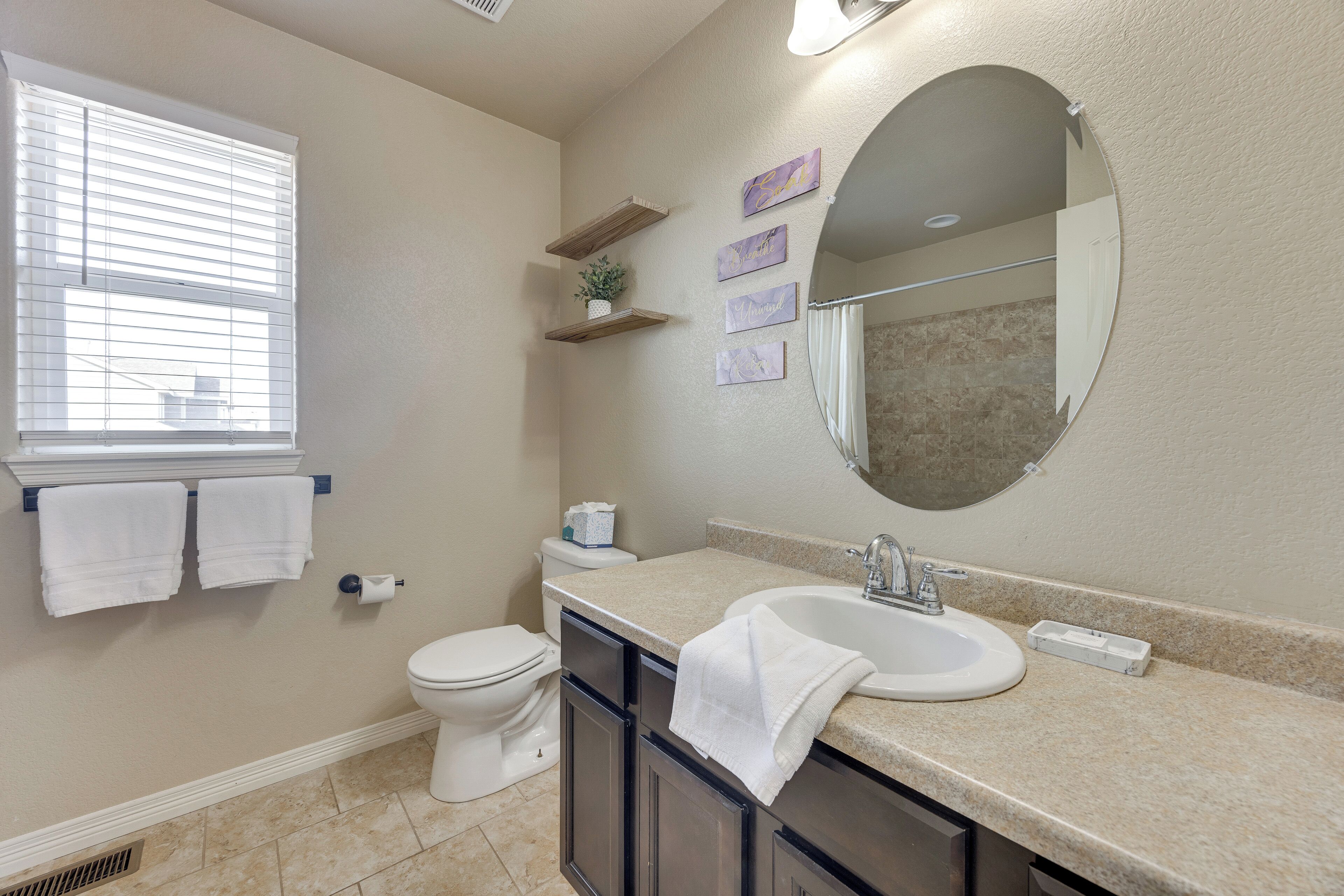 Une salle de bain avec des toilettes, un lavabo et un miroir. Des serviettes sont accrochées au mur et il y a une fenêtre avec des stores.