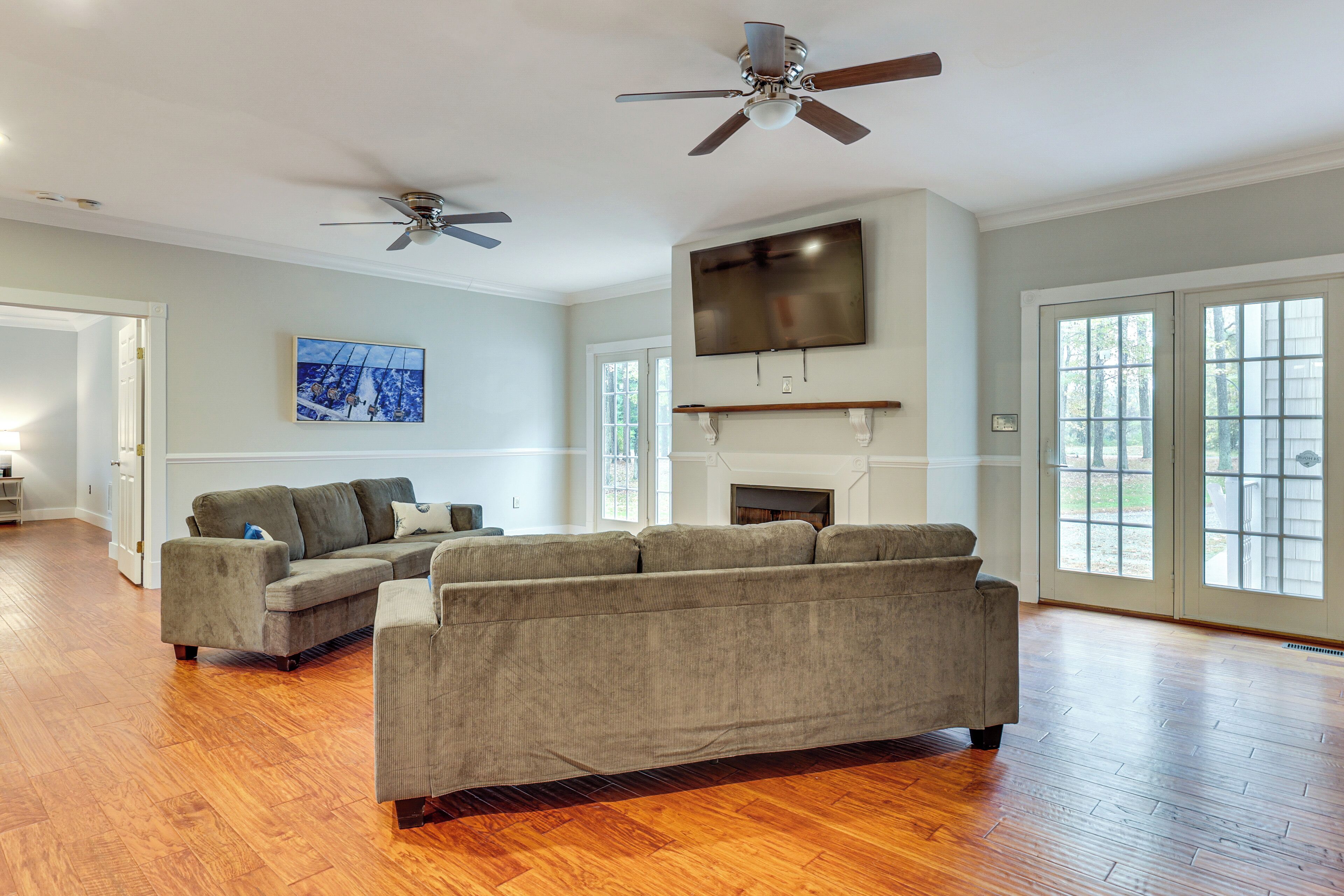 A spacious living room with hardwood flooring, a large sectional sofa, a flat-screen TV mounted above a fireplace, and two ceiling fans.