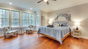 A bedroom with a large bed, two upholstered chairs, a small table, a ceiling fan, and a large window with a view of trees. - Cow Shed Waterfront 3BR Home w/ Boat Dock! (Lancaster)