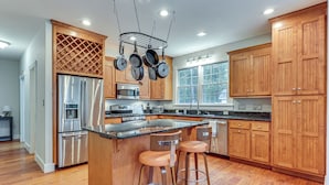 A modern kitchen with wooden cabinets, stainless steel appliances, and a central island with bar stools. - Cow Shed Waterfront 3BR Home w/ Boat Dock! (Lancaster)