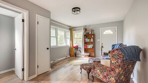 Una habitación con suelo de madera, un sillón floral, una estantería y una puerta que da a otra estancia.