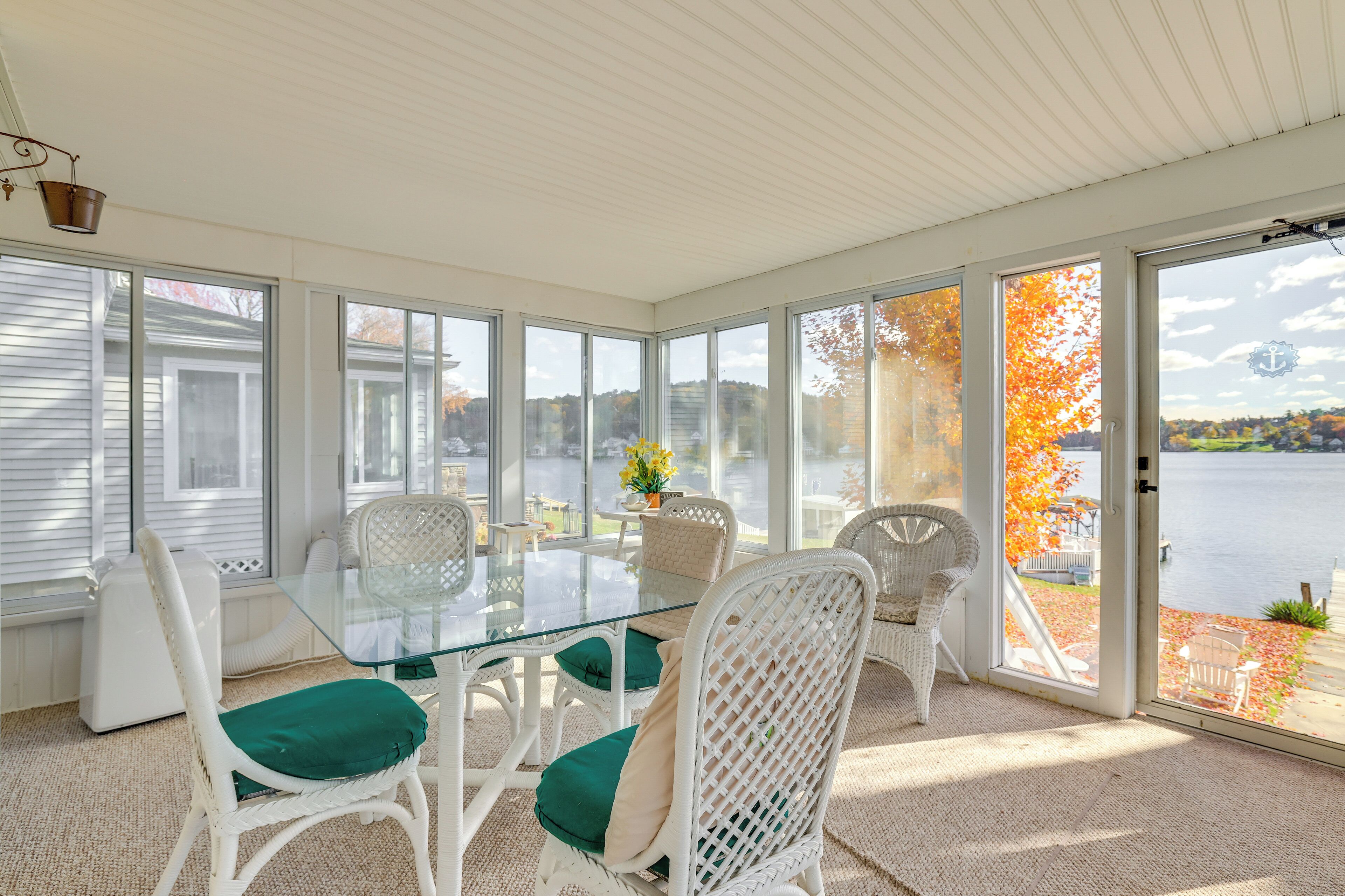A sunroom with a glass table and white wicker chairs, large windows, and a view of a lake.