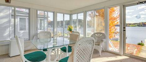 A sunroom with a glass table and white wicker chairs, large windows, and a view of a lake.