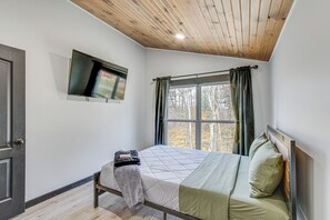 A bedroom with a bed, a TV mounted on the wall, a window with curtains, and a wooden ceiling. - 'chène Cottage' Near Burr Oak Lake & Trailheads! (Glouster)