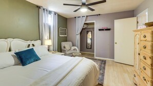 Un dormitorio con cama, una silla, cómoda, ventilador de techo y puerta que da a otra habitación.