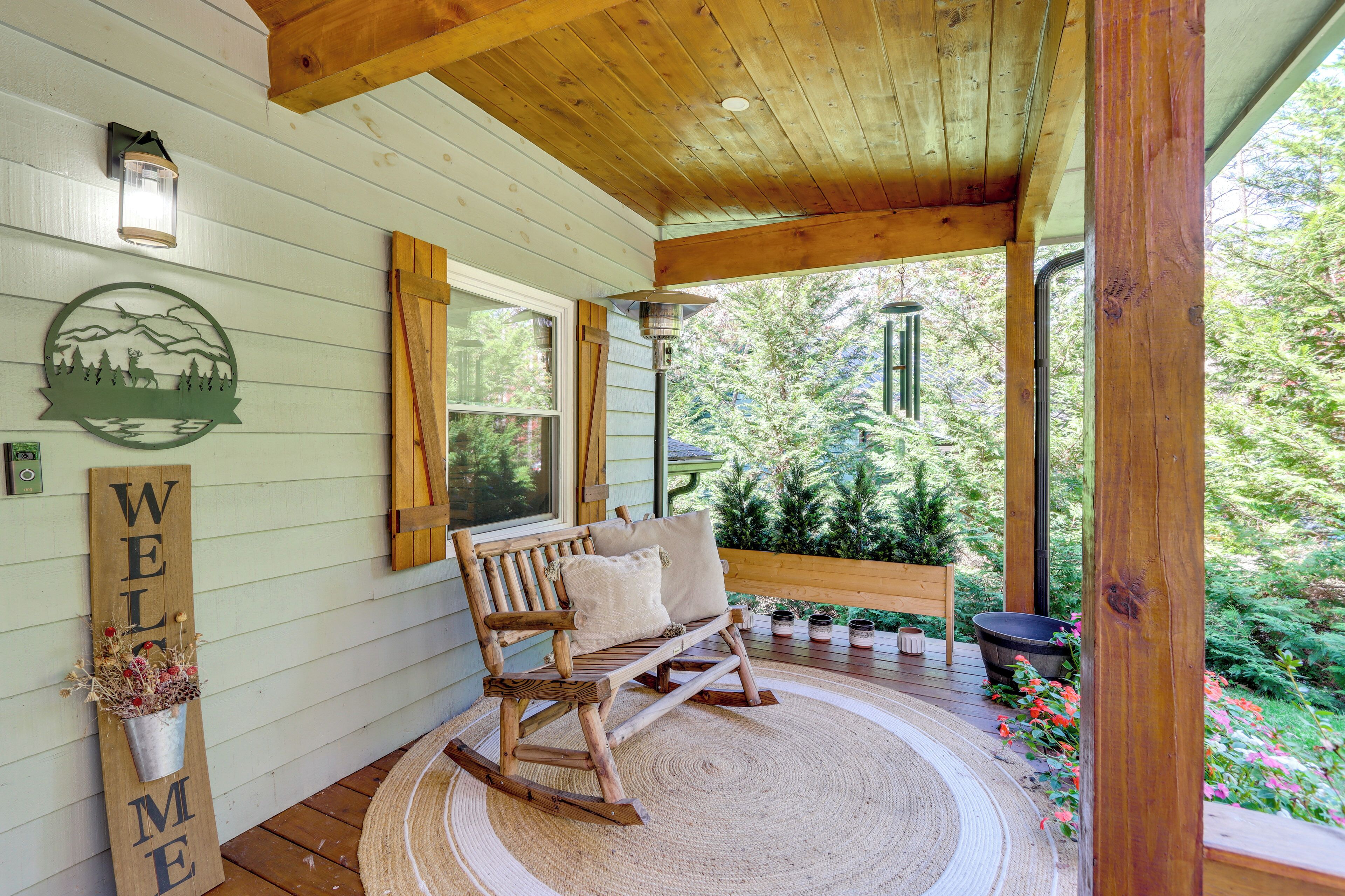 Une véranda en bois avec une chaise berçante, une pancarte de bienvenue et une vue sur un jardin.