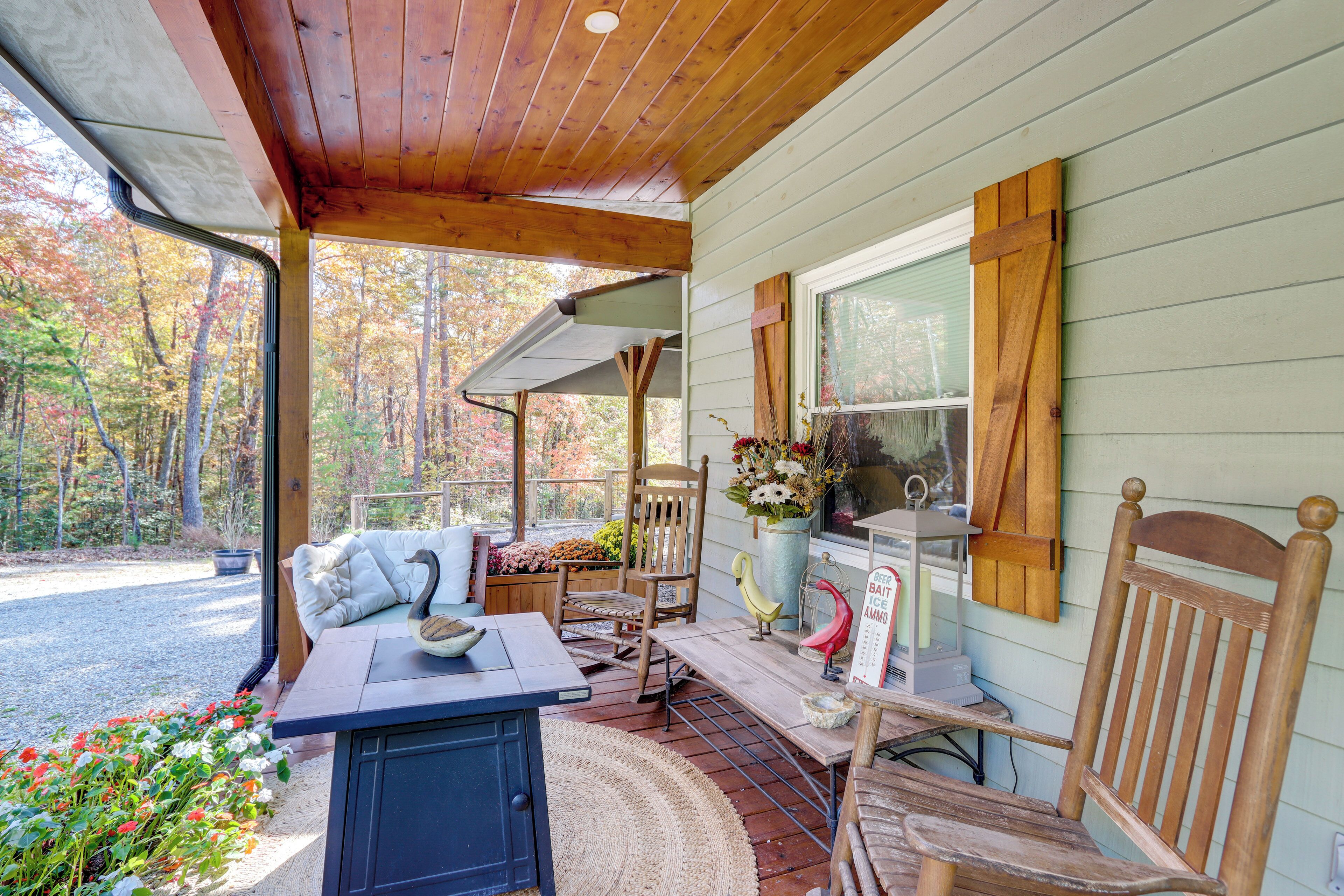 A covered porch with wooden furniture, a table, and chairs. There is a window with a view of trees and a potted plant on the table.
