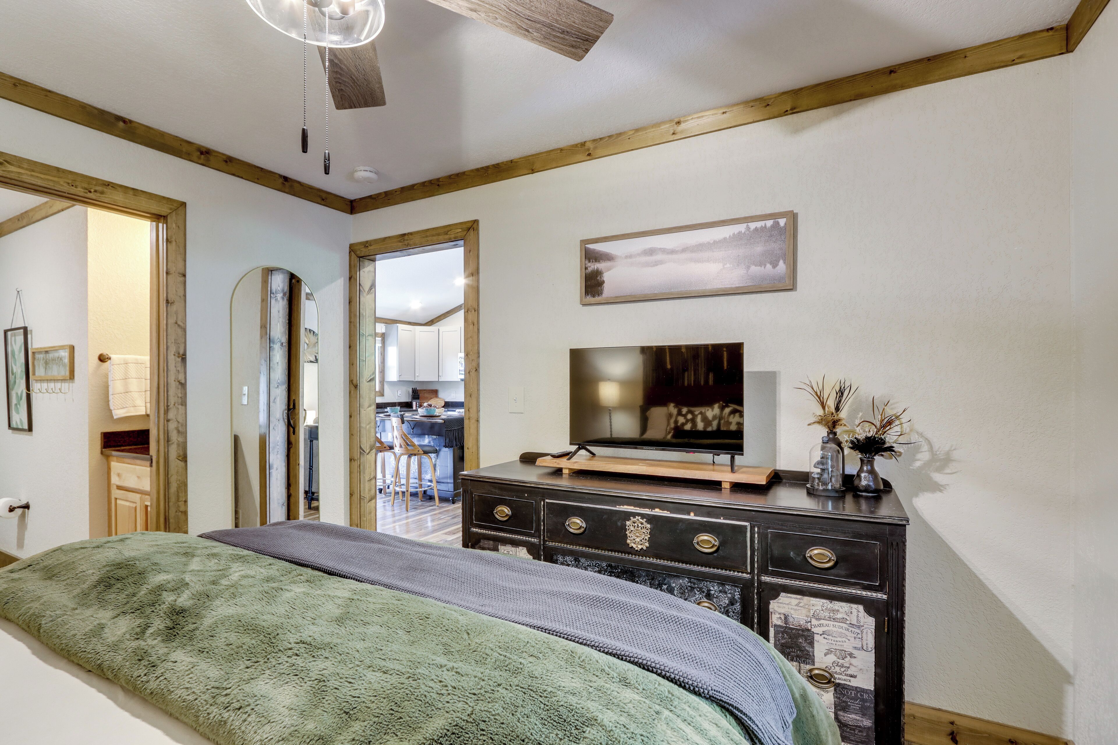 A bedroom with a bed, dresser, TV, and a view of the kitchen.