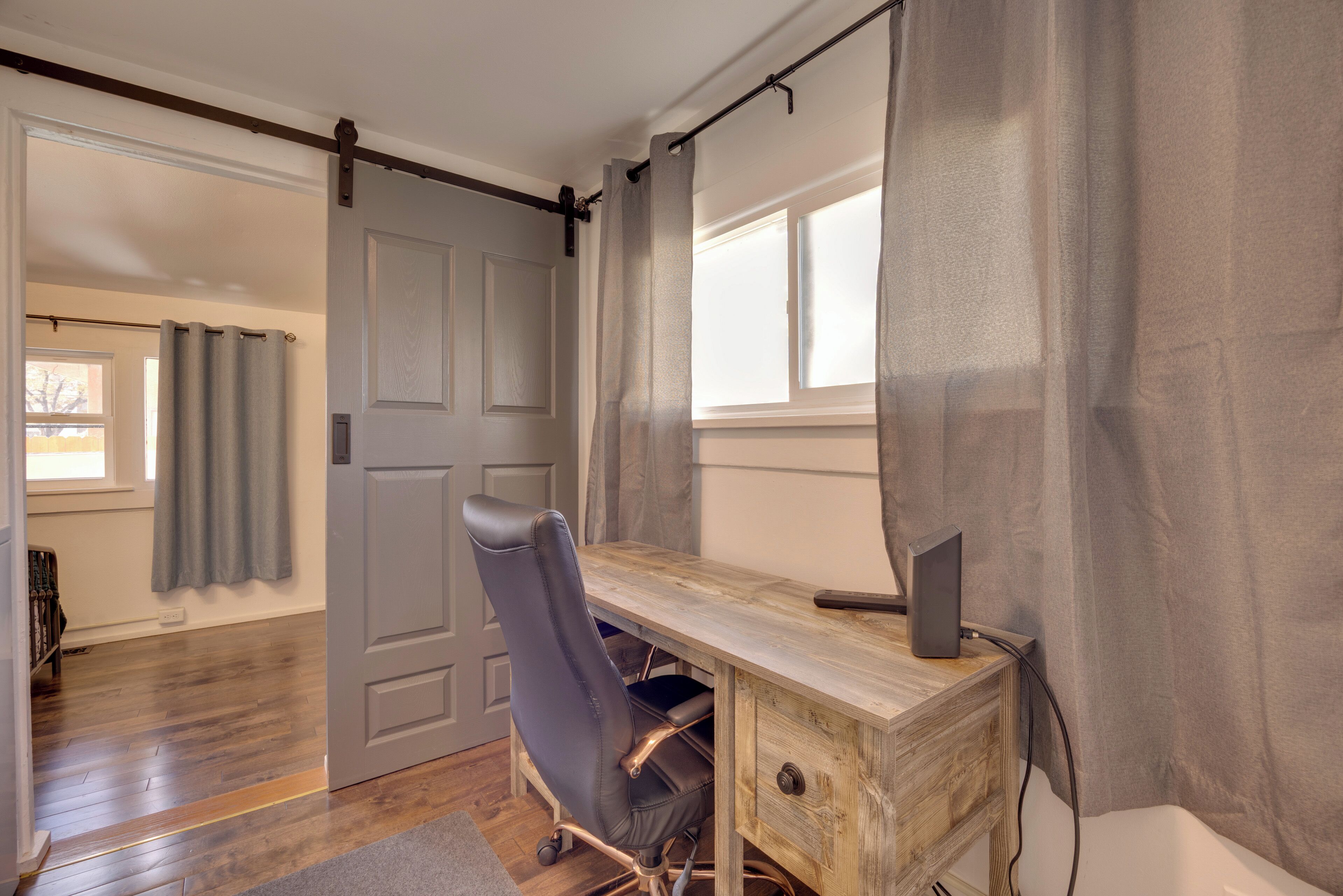 A room with a wooden desk, a black office chair, a sliding door, and a window with curtains.
