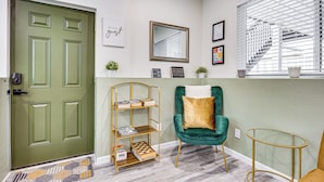 Un pasillo con una puerta verde, una estantería, un sillón verde, una mesa de vidrio y una ventana con persianas.