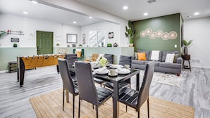 Una sala de estar moderna con una mesa de fútbol de mesa, una zona de comedor con una mesa dispuesta para cuatro personas y un sofá gris con cojines anaranjados.