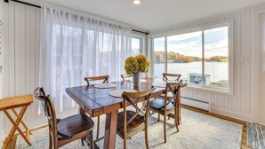 Ein Esszimmer mit Holztisch und -stühlen, einer Vase mit gelben Blumen und Blick auf ein Gewässer und Bäume durch große Fenster.