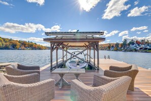Une terrasse en bois, agrémentée de meubles en osier et d’une table, qui offre une vue sur un lac, avec des maisons en arrière-plan.
