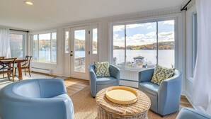 Ein geräumiges Zimmer mit großen Fenstern und Blick auf einen See und Herbstlaub. Zwei blaue Sessel und ein Korbtisch sind vorhanden.