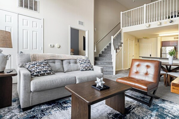 A living room with a grey sofa, a brown leather chair, a wooden coffee table, and a staircase leading to an upper level. - Landing Apartments New Haven (New Haven)