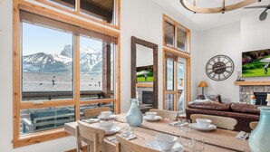 A dining area with a wooden table set for four, large windows with a mountain view, a brown leather sofa, and a wall clock. - Majestic Views! Tamarack Penthouse 3 BD / 3 BA (Canmore)