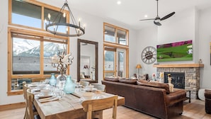A spacious living area with a fireplace, a dining table set for six, and a large window offering a mountain view. - Majestic Views! Tamarack Penthouse 3 BD / 3 BA (Canmore)