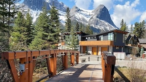 A wooden bridge leading to a modern building with large windows, surrounded by snow-capped mountains and evergreen trees. - Majestic Views! Tamarack Penthouse 3 BD / 3 BA (Canmore)