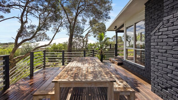 Um deck de madeira com mesa e bancos, cercado por uma parede de tijolos pretos e grandes janelas.