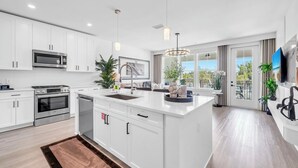 A modern kitchen with white cabinetry, a central island, stainless steel appliances, and pendant lighting. - The Grove Holly 125 - 1 Bedroom - Lazy River - Seascape Resort (Miramar Beach)
