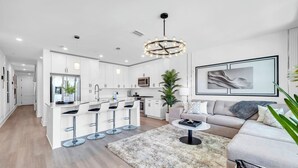 A modern kitchen with white cabinets, a central island with bar stools, a dining area with a sofa and coffee table, and a large abstract painting on the wall. - The Grove Holly 125 - 1 Bedroom - Lazy River - Seascape Resort (Miramar Beach)