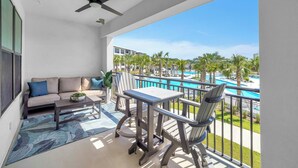 A balcony with a sofa, table, chairs, and a view of a pool and palm trees. - The Grove Holly 125 - 1 Bedroom - Lazy River - Seascape Resort (Miramar Beach)