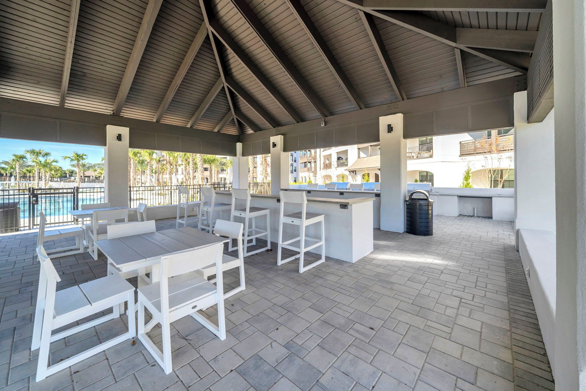 An outdoor patio with white furniture, a bar area, and a view of a pool and buildings in the background.