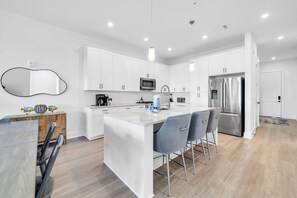 A modern kitchen with white cabinets, stainless steel appliances, and a central island with bar stools. - The Grove Juniper 243 - 2 Bedrooms - Lazy River - Seascape Resort (Miramar Beach)